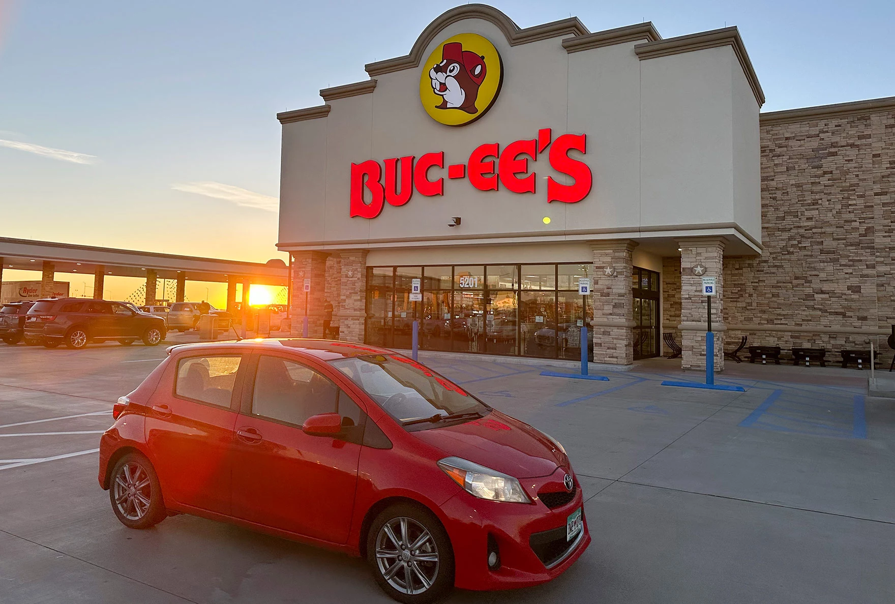 Red compact car parked in front of Buc-ee's store at sunset.